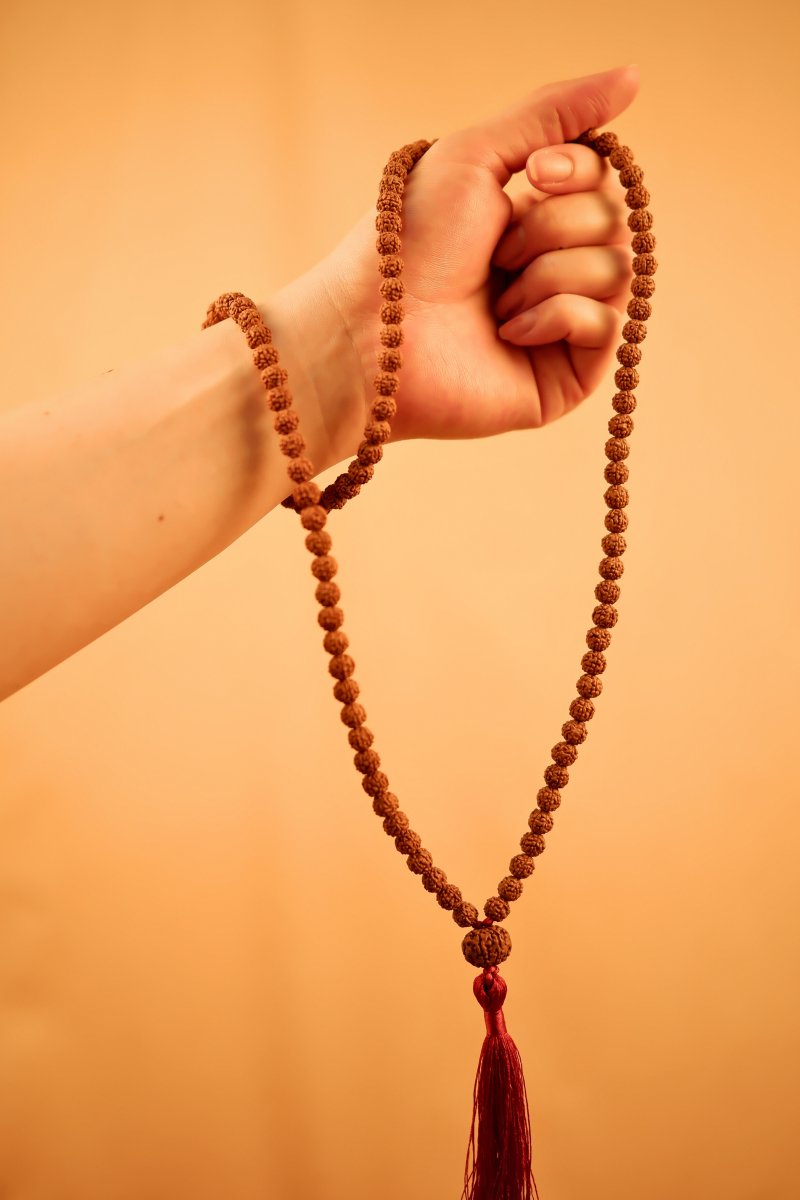 Hand holding a sacred Java Rudraksha mala with red tassel on warm beige background.